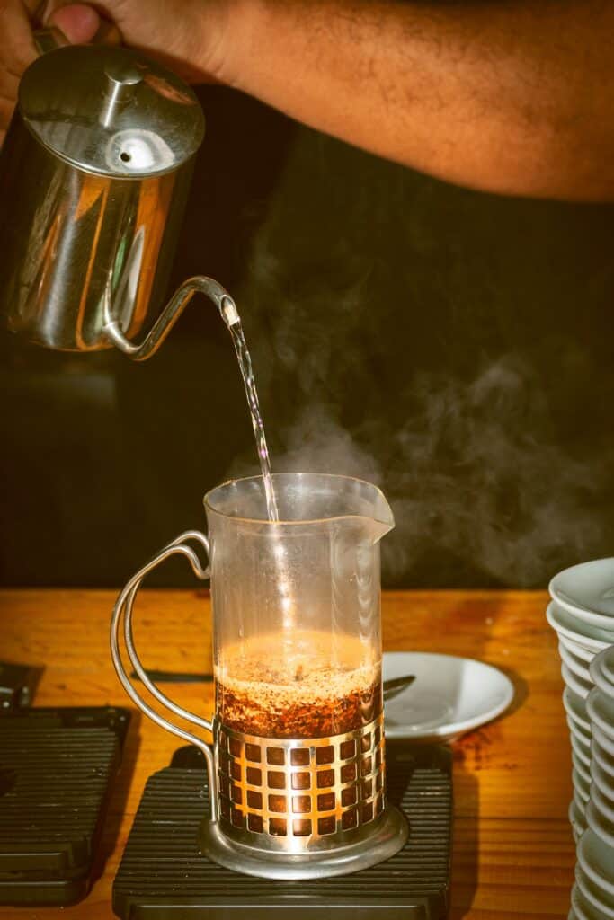 someone pours hot water from a metal pot into a glass french press coffee maker