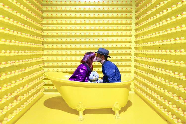 two people kiss inside a yellow bathtub in a yellow room in a museum art exhibit