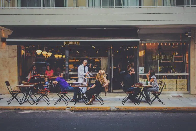 diners sit outside a restaurant while a server talks to one of the patrons
