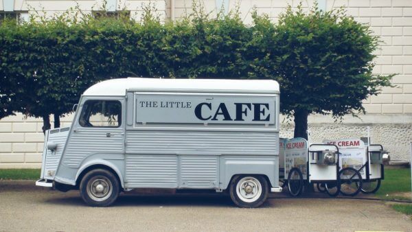 a silver truck with a sign reading 'the little cafe' is parked in front of a row of trees
