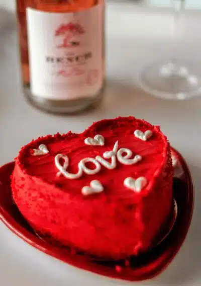 the word 'love' is written in white icing on a red velvet heart cake with a bottle of rosé wine pictured in the background