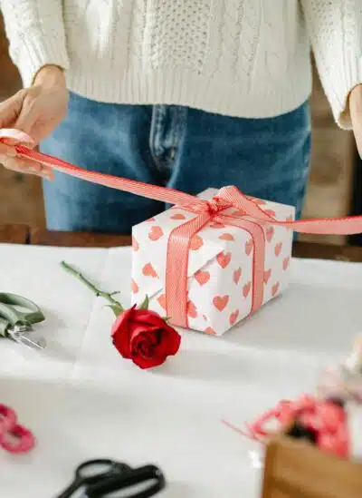 a retail employee wraps a git box in pink heart paper and a pink bow, while a red rose sits on the table