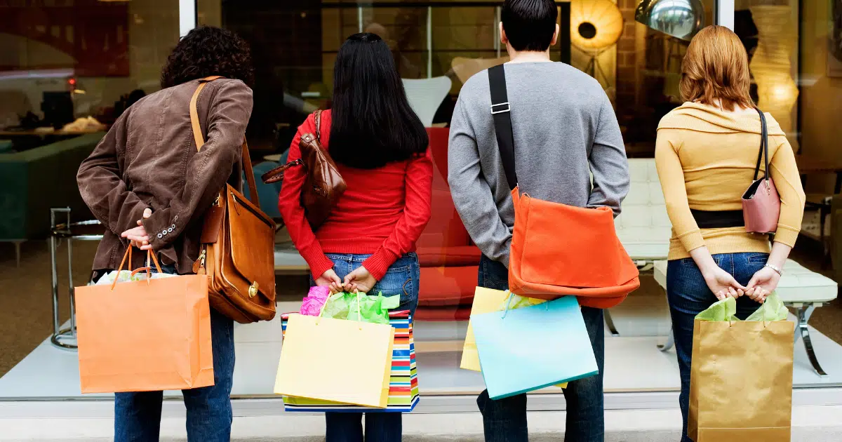 shoppers holding bags stand outside of a furniture store and look through the window