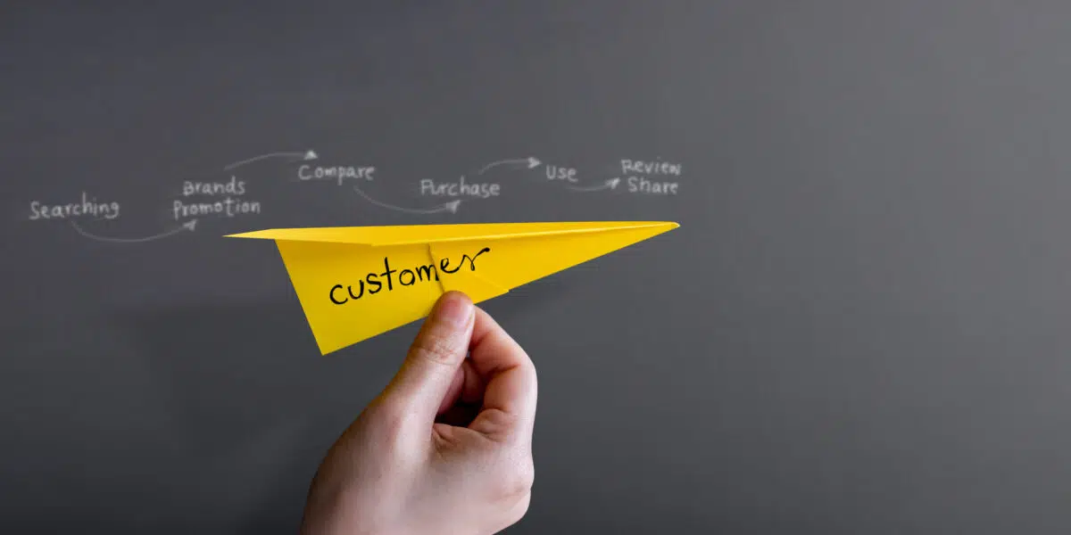 a person holds a yellow paper airplane that reads customer with a purchase flow chart above it