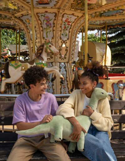 two kids sit in front of a merry go round holding a dinosaur stuffed animal prize 