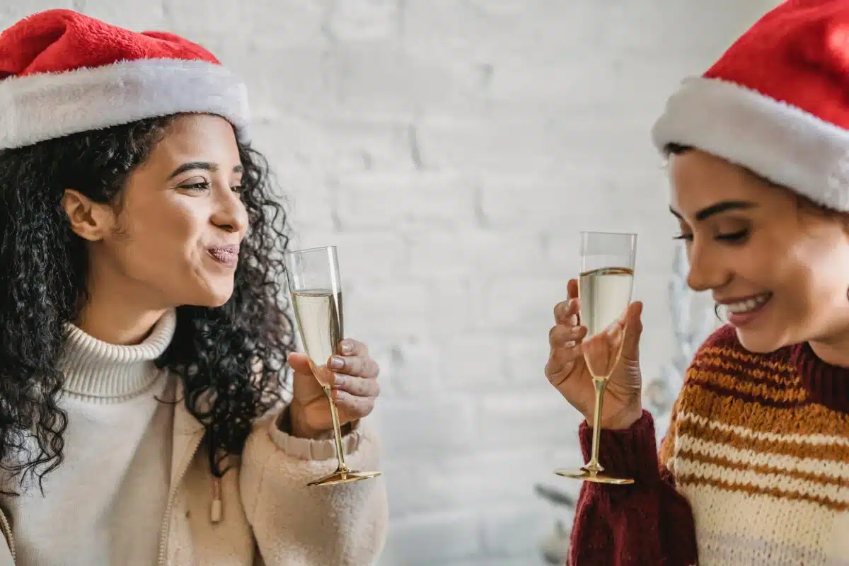 two friends in Santa caps smile as they drink champagne