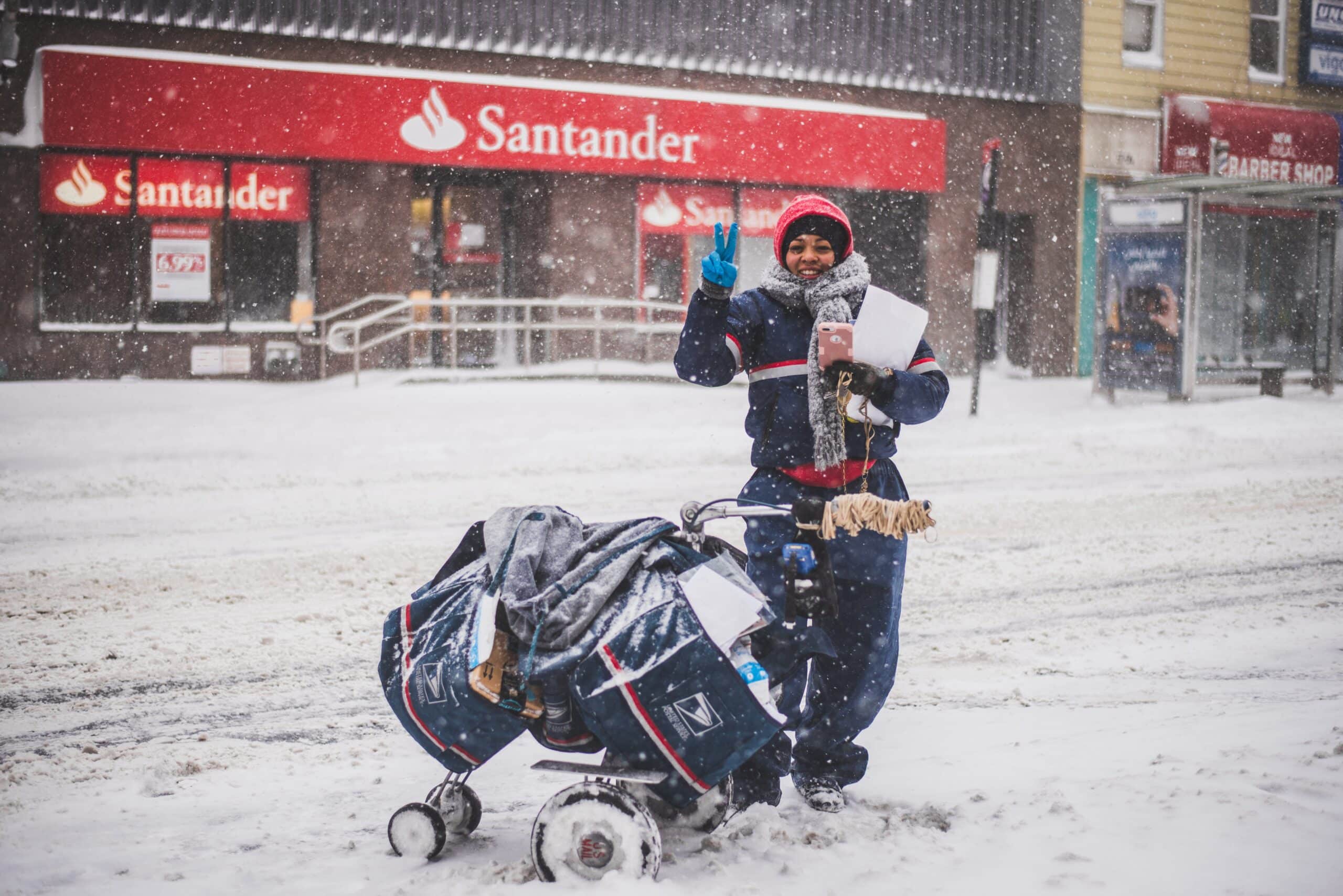 a USPS worker waves a peace sign while pushing a mail cart through the snow