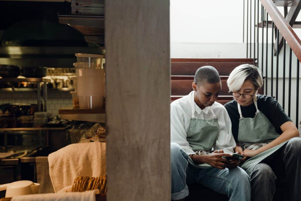 two workers sit on a staircase and look over a retail employee scheduling software app on their smartphone