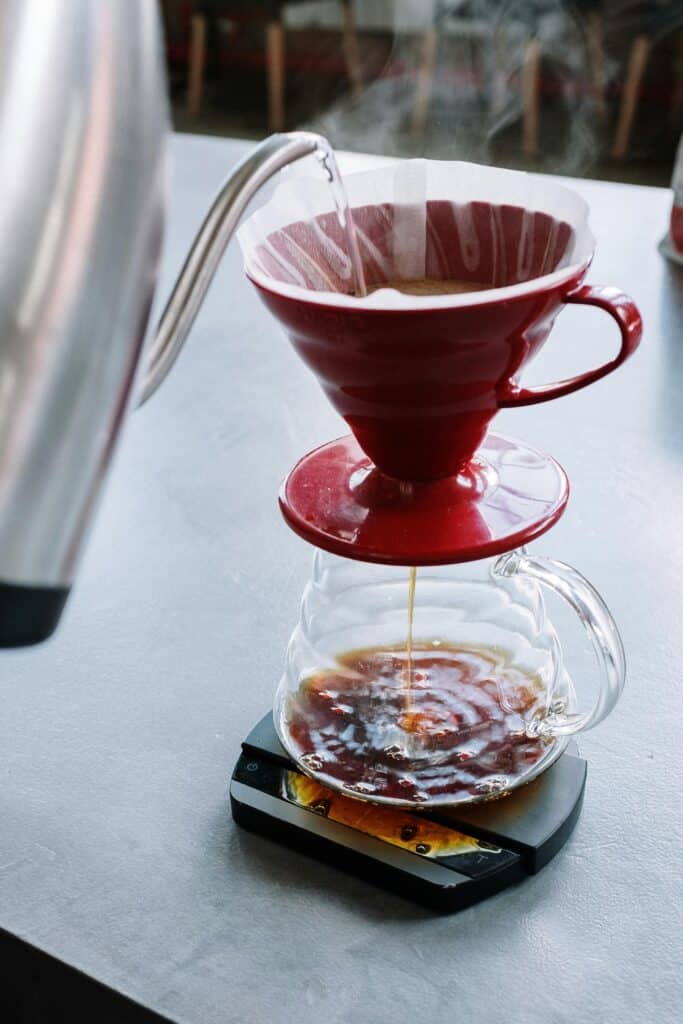 hot water is poured from a gooseneck kettle through a red ceramic cone and into  glass pitcher